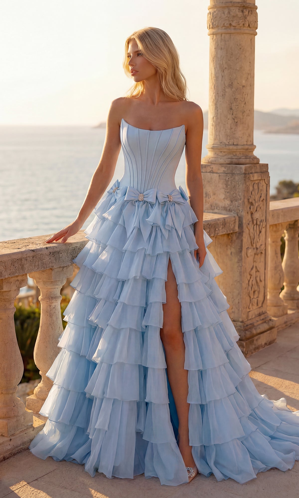 A woman in the PromGirl 250071 light blue Tiered Prom Ball Gown with Bows stands on a stone balcony overlooking the sea at sunset.